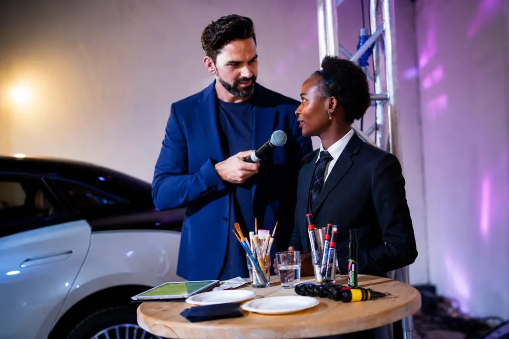 Professional event MC in Dubai interviewing a guest during a live corporate activation, engaging the audience with an interactive microphone session beside a branded display.