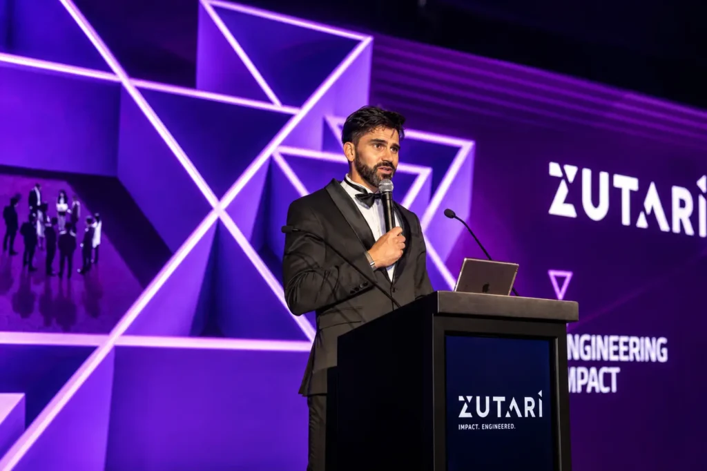 Event host presenting from a podium while addressing attendees at a corporate conference, with illuminated geometric stage design, branded visuals and professional event production.